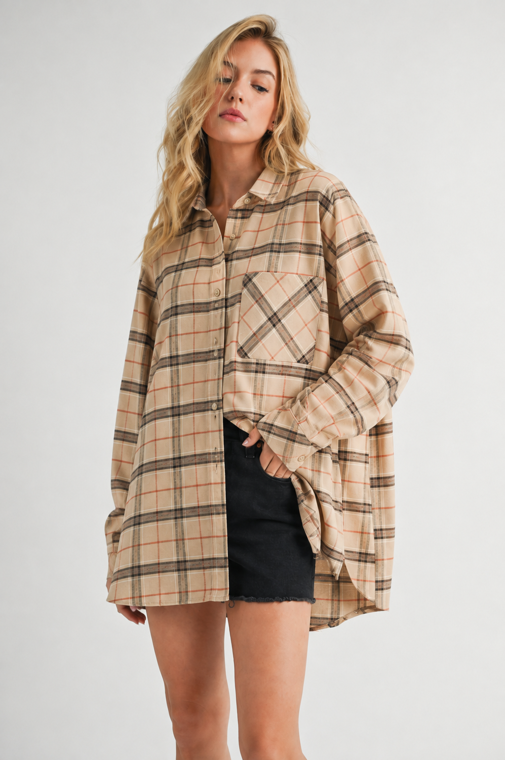 A woman modeling an oversized beige plaid button-up shirt with black shorts against a light gray studio background.