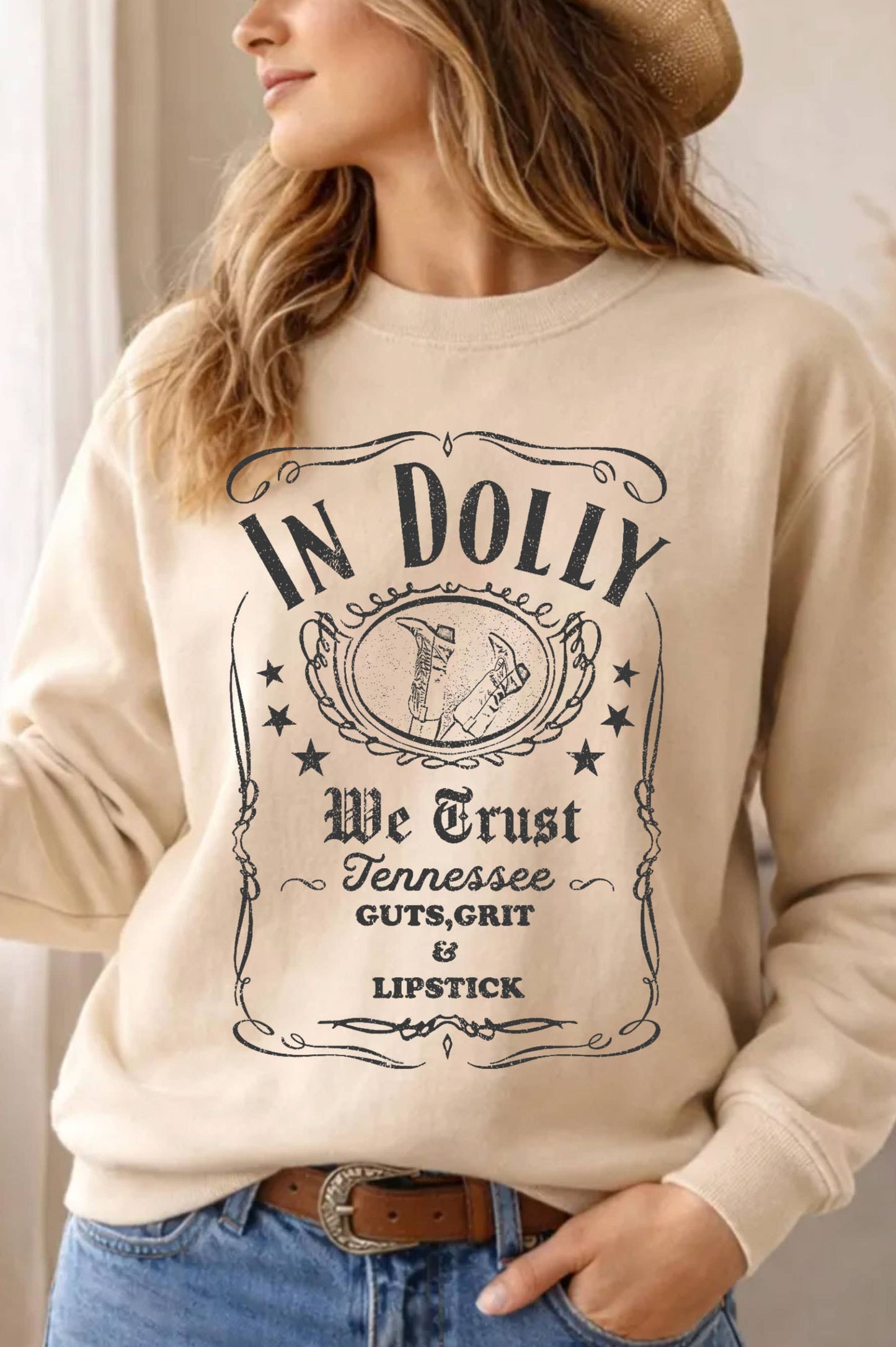 A woman wearing a beige crewneck sweatshirt with a vintage-style black graphic print that reads 'IN DOLLY We Trust Tennessee GUTS, GRIT & LIPSTICK'.