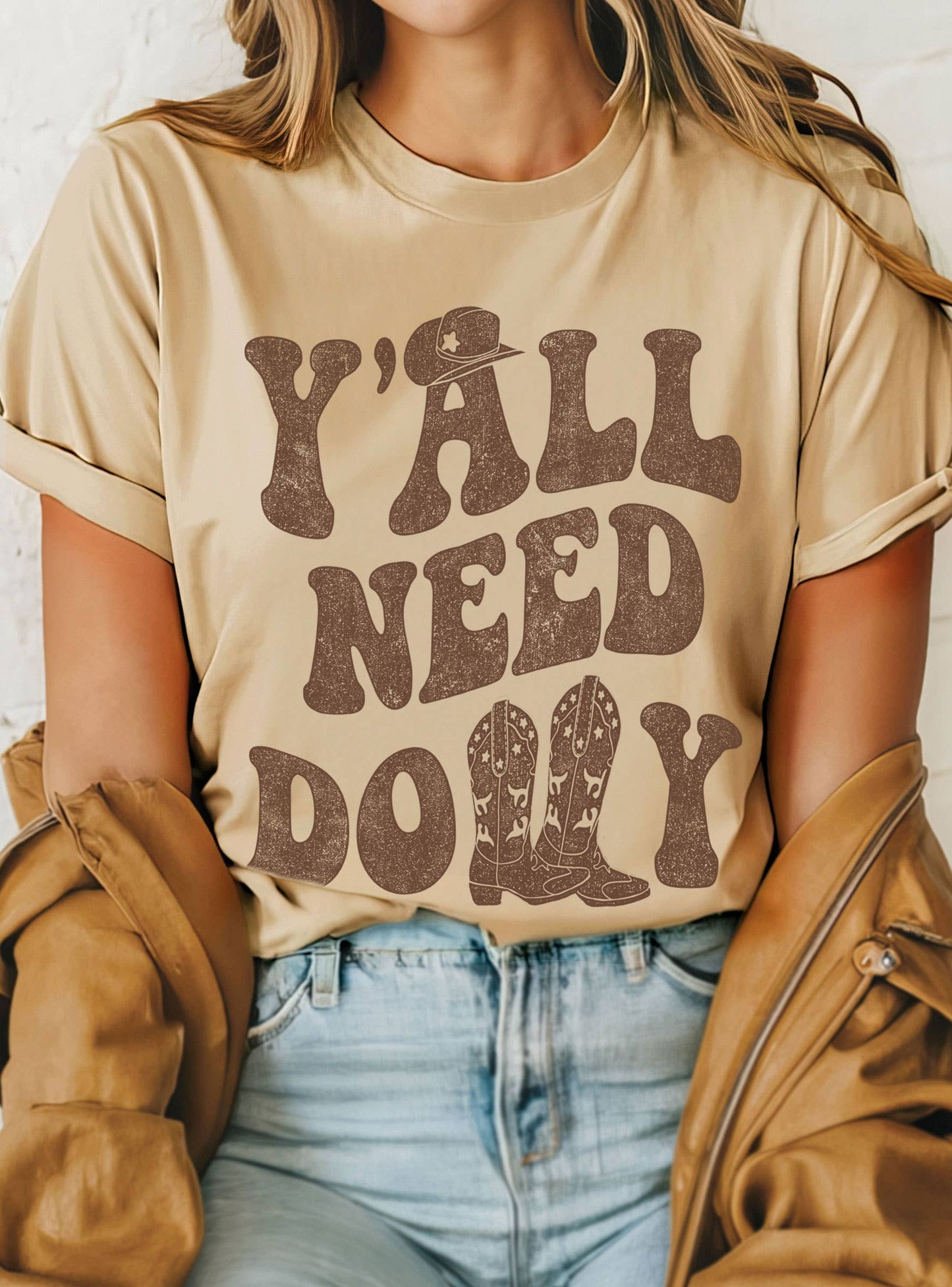 Person wearing a beige t-shirt with 'Y'all Need Dolly' text, sitting against a white brick wall.